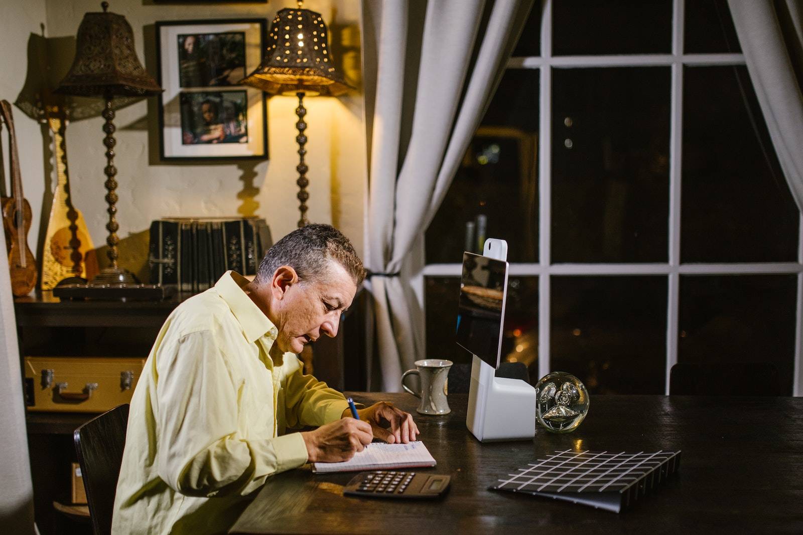 Side view of concentrated mature male accountant in shirt taking notes in planner while sitting at table in classic styled apartment and working remotely using calculator and tablet